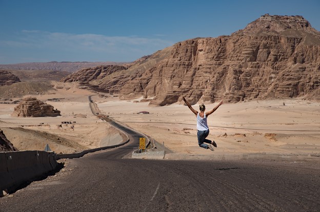 A Woman Jumping on Road With Desert Land on Either Sides